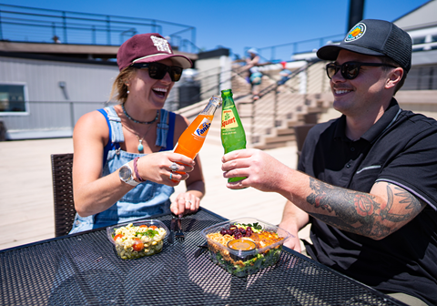 Woman and man sitting outside clinking soda bottles with pre packaged salads