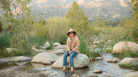 Singer Songwriter Brett Dennen sitting on a rock in a creek.