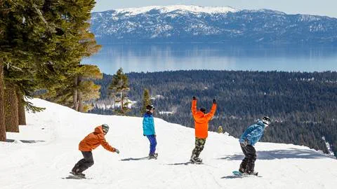 Four snowboarders going down a flat run in front of a perfect view of Lake Tahoe.