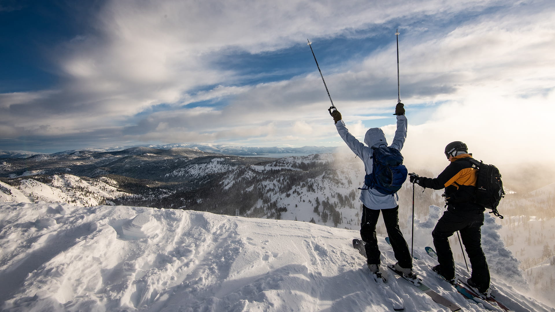 Two skiers, one with poles raised towards the sky, atop a summit at Alpine in Lake Tahoe.