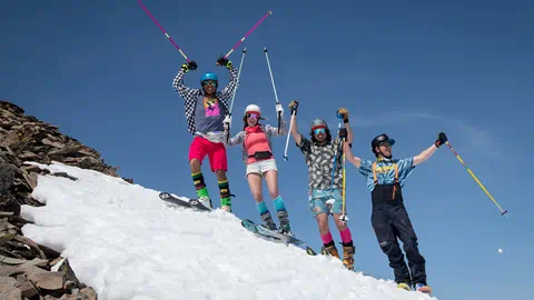 Skiers dressed in short shorts and sunglasses stand on top of a peak with their poles up in Spring.