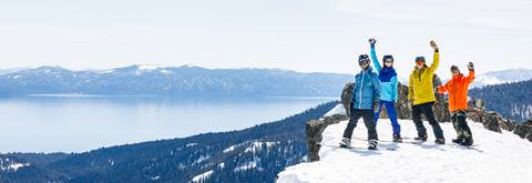 4 Snowboarders smiling and waving with Lake Tahoe in the background