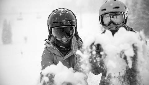 Two young women dressed in winter sports gear hold out their snow-filled hands toward the camera. 