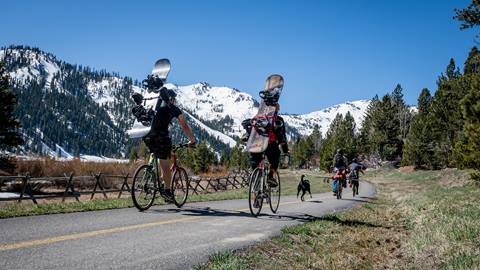 People riding bikes along the Olympic Valley bike path with snowboards heading to The Spring Skiing Capital