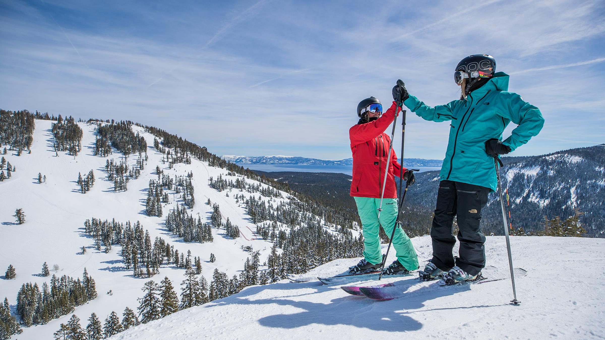 two female skiers high five at the top of a run at Alpine Meadows with lake tahoe in the background