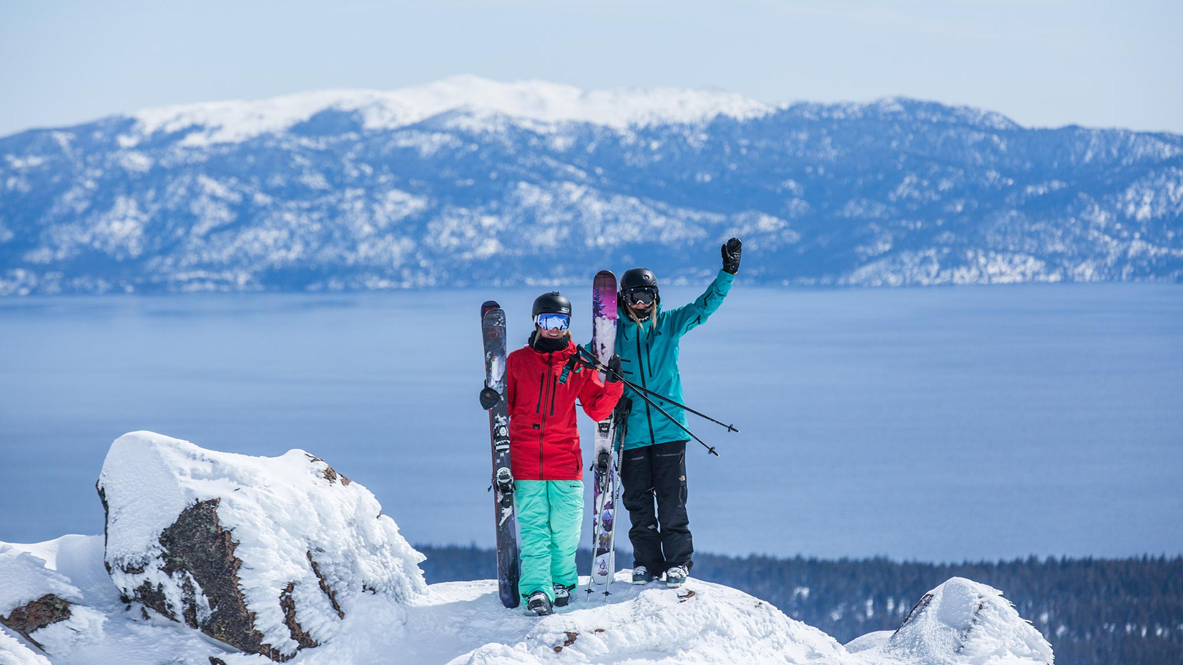 Two girls wave from the top of a peak at Alpine Meadows with Lake Tahoe in the background